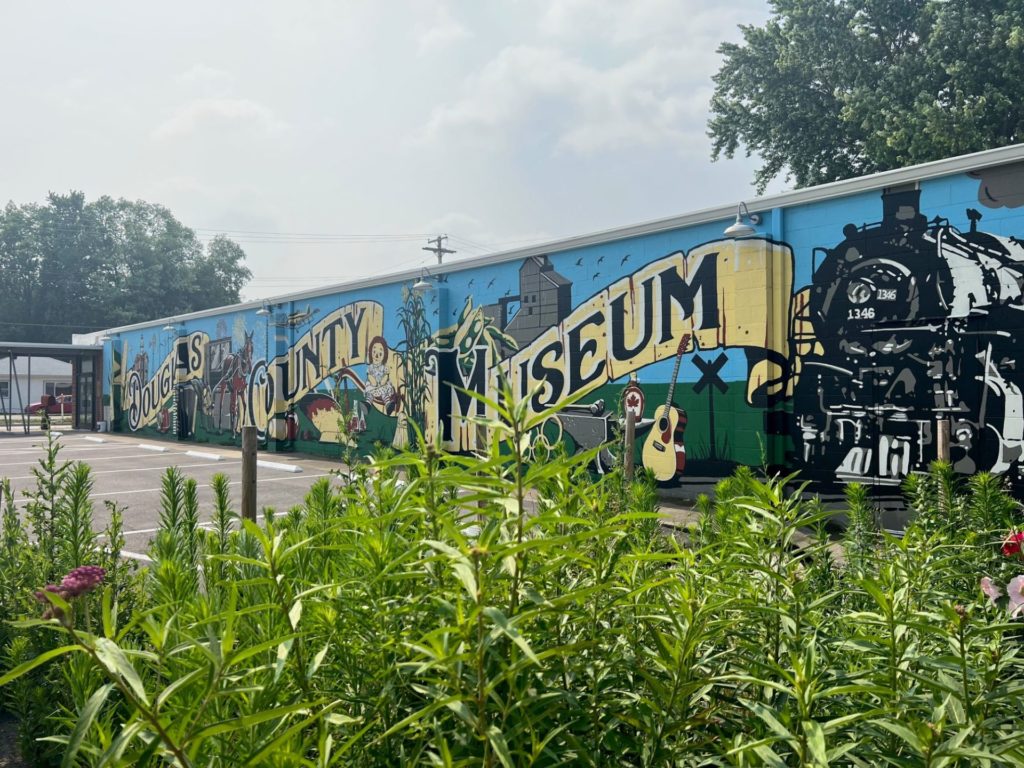 A long one story building with a large colorful mural painted along the side. There are yellow painted banners that say Douglas County Museum in black lettering. In the foreground is a patch of tall plants and flowers.