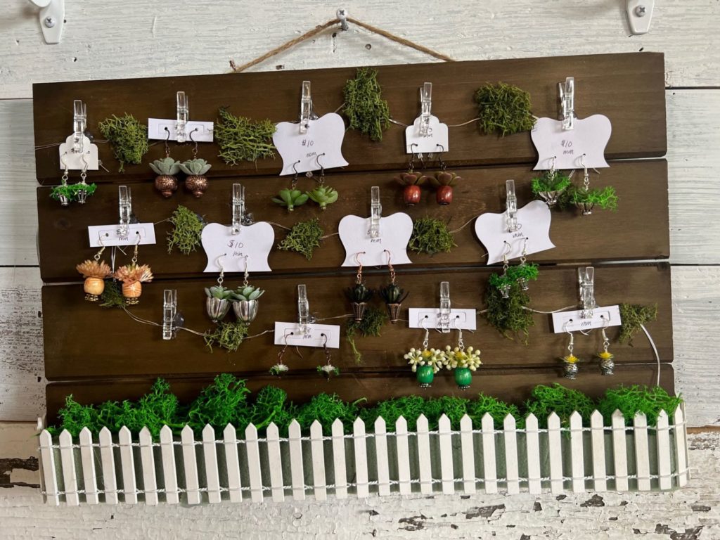 A disply with wood planks and a white picket fence. Earrings in the shape of potted plants are hanging on the display.