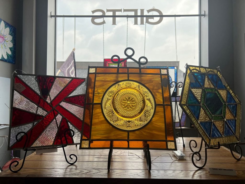Three stained glass tiles on black metal display easels are sitting in front of a window on a wooden shelf.