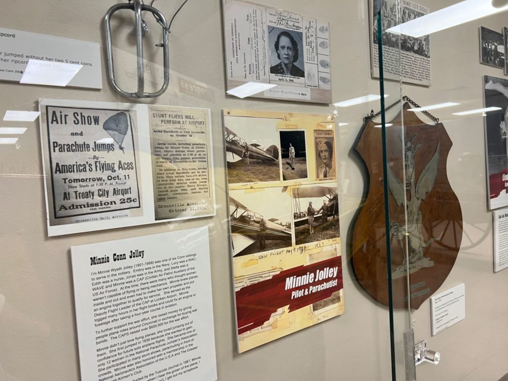 A museum display behind glass with informational panels and a photo panel with the words Minnie Jolley, Pilot & Parachutist in white lettering.