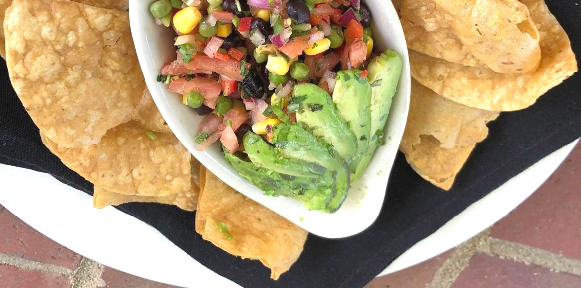 A cropped photo of vegan caviar with avocado slices, corn, black beans, and tomatoes in a white boat on a plate with tortilla chips on a black napkin for the Vegan Chef Challenge Champaign. Photo by Silvercreek Restaurant on Instagram.