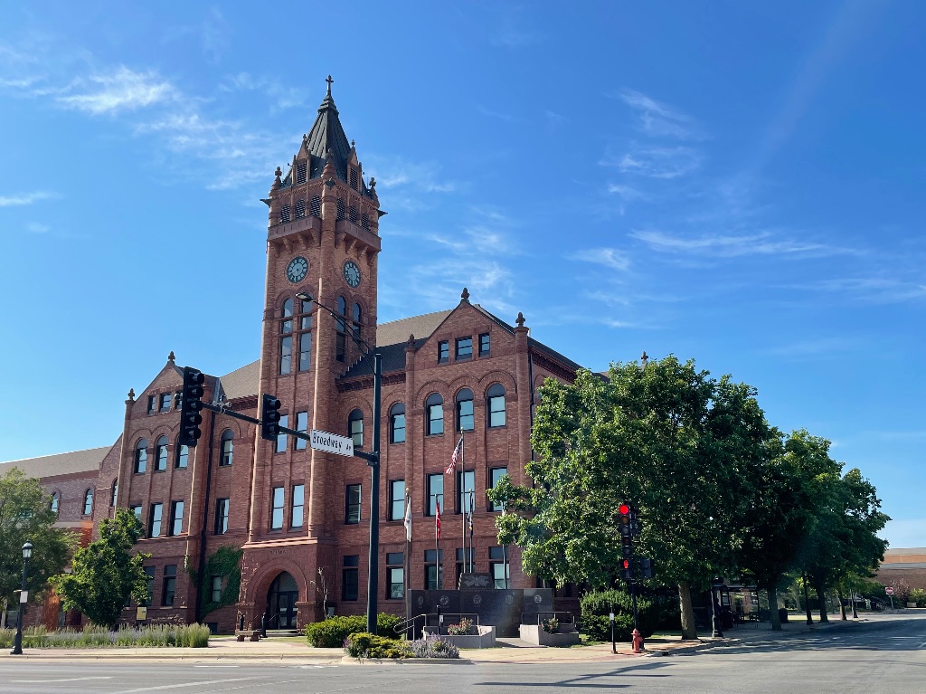 A brown brick building with a clock tower in the center, sits on a corner surrounded by green leafy trees. The sky is blue with a few whispy clouds behind. 