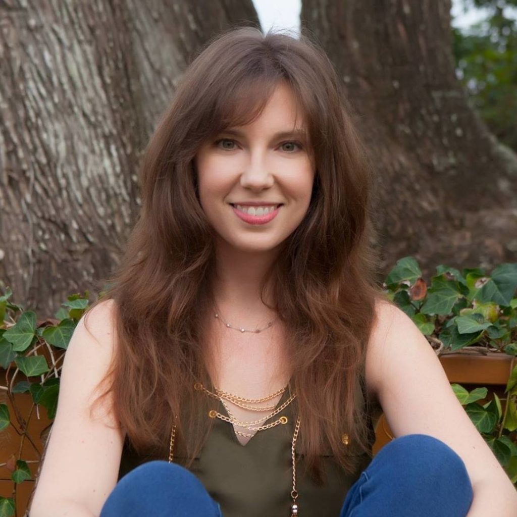 Christina Delay sits on the ground outside in front of a tree with her legs crossed. She is white, with long brown hair with bangs. She wears jeans and a green sleeveless top while smiling at the camera. 