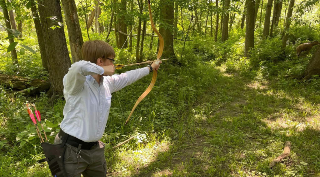A white person wearing a light colored button up shirt in the woods is aiming a bow and arrow at a fake deer target. They have a sleeve with some red-feathered arrows on their hip.