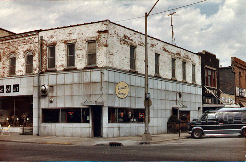 A street corner with a sort of dirty white brick and gray paneled facade. There is a circular yellow sign that says Esquire Lounge in script.