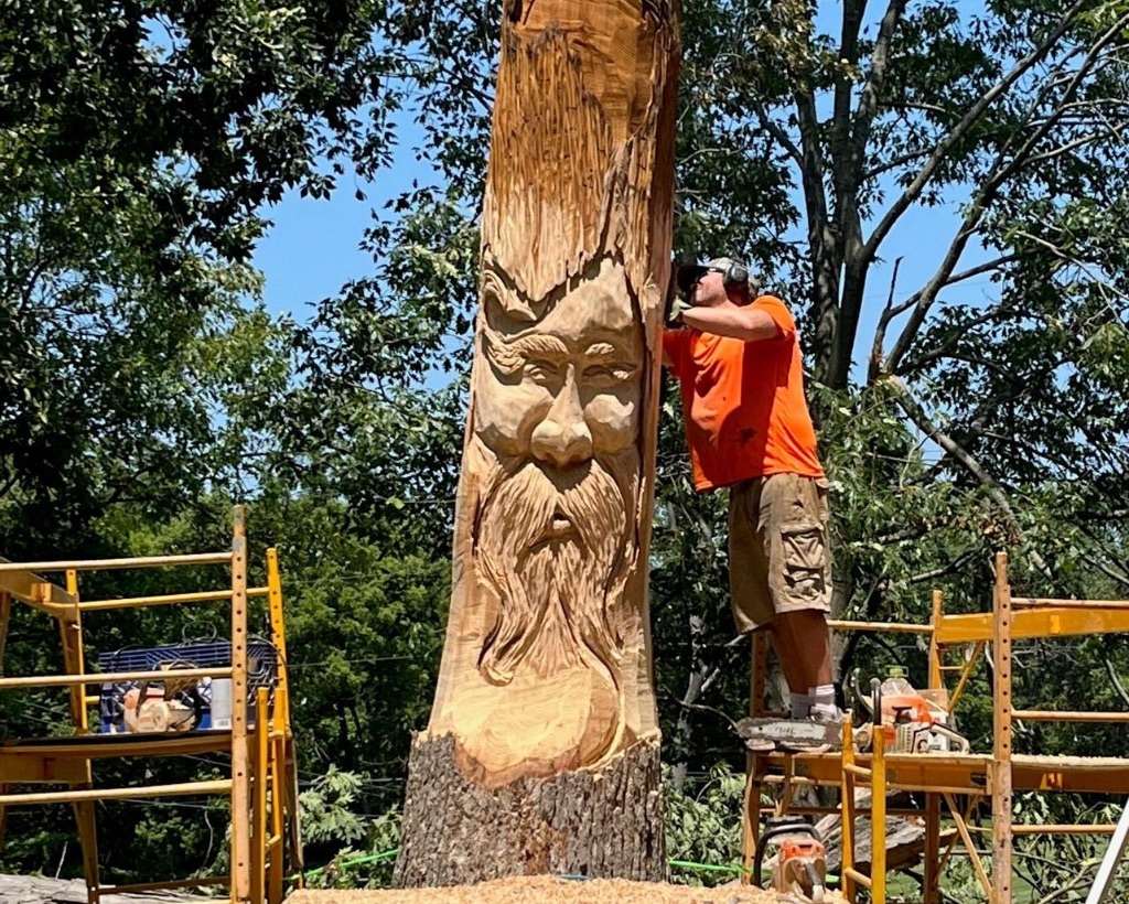 A man stands next to a dead tree with a chainsaw carving a face into the trunk.