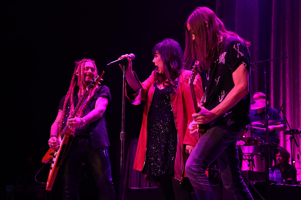 This is a photo of a rock band performing on a stage. The band consists of three members, two guitarists and a drummer. The guitarist on the left is wearing a black leather jacket and a black hat, while the guitarist on the right is wearing a black t-shirt and has long hair. The drummer is in the background and is not clearly visible. The stage is lit with purple and red lights.