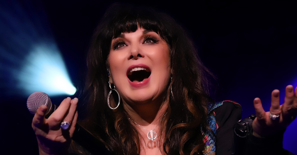 This is a photo realistic image of a person singing on stage with a microphone in hand. The person is wearing a black top with a colorful pattern on it and a silver necklace. The person is also wearing silver hoop earrings. The background consists of blue stage lights.