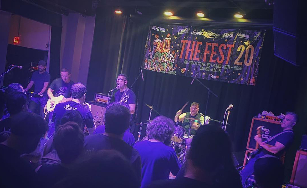 This is a photo of a band performing on a stage in a dark venue. The stage has a large banner that reads “THE FEST 20” in colorful letters. The band members are playing various instruments, including guitars, drums, and a bass. The audience is visible in the foreground, with their backs to the camera. The lighting is dim, with purple and blue lights illuminating the stage.