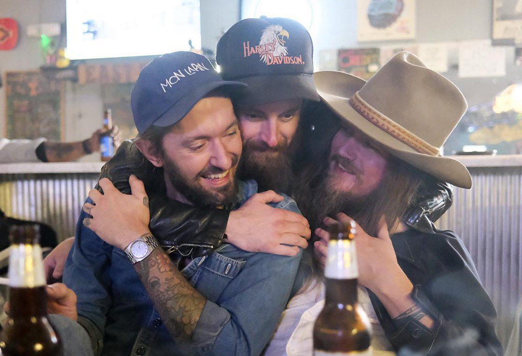 A man in a Harley Davidson hat behind two other men with hats on putting his arms around them in an embrace.