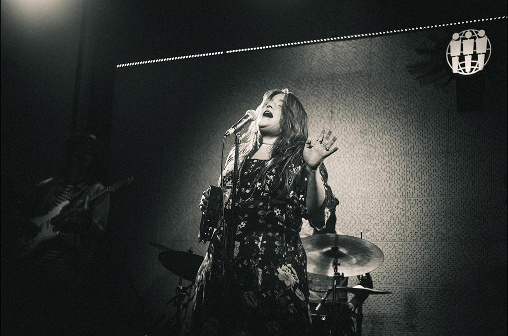 A black and white photo of a woman with her eyes closed, holding a tambourine, leaning back and singing into a microphone.