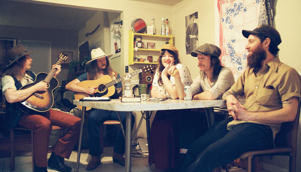 5 members of Banditos around a kitchen table. Two members are playing acoustic guitar, and everyone has a hat on.