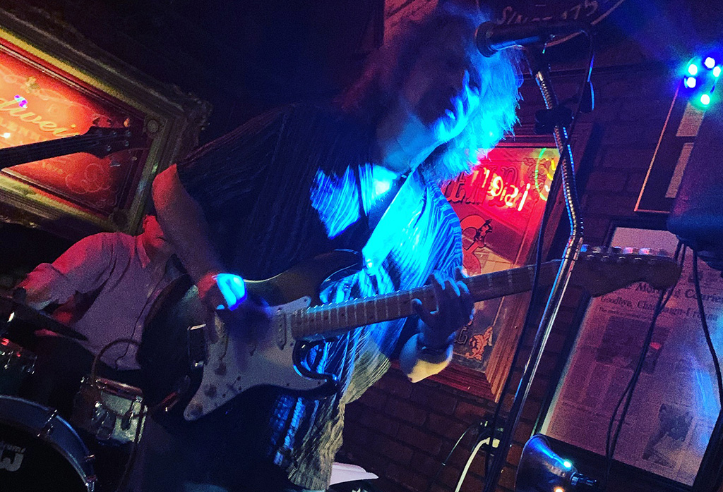 Main playing guitar on a dimly lit stage with a drummer behind him