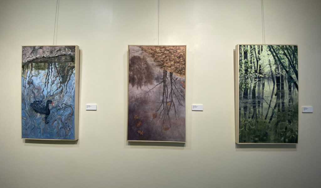 CIA Show at Springer Cultural Center; three large vertical rectangular paintings hang on a gallery wall. The left one is primarily blue and features a black swan in a pond; the middle painting is mostly mauve and shows a tree reflecting in a puddle, the right painting is mostly green and shows a forrest reflecting in water.
