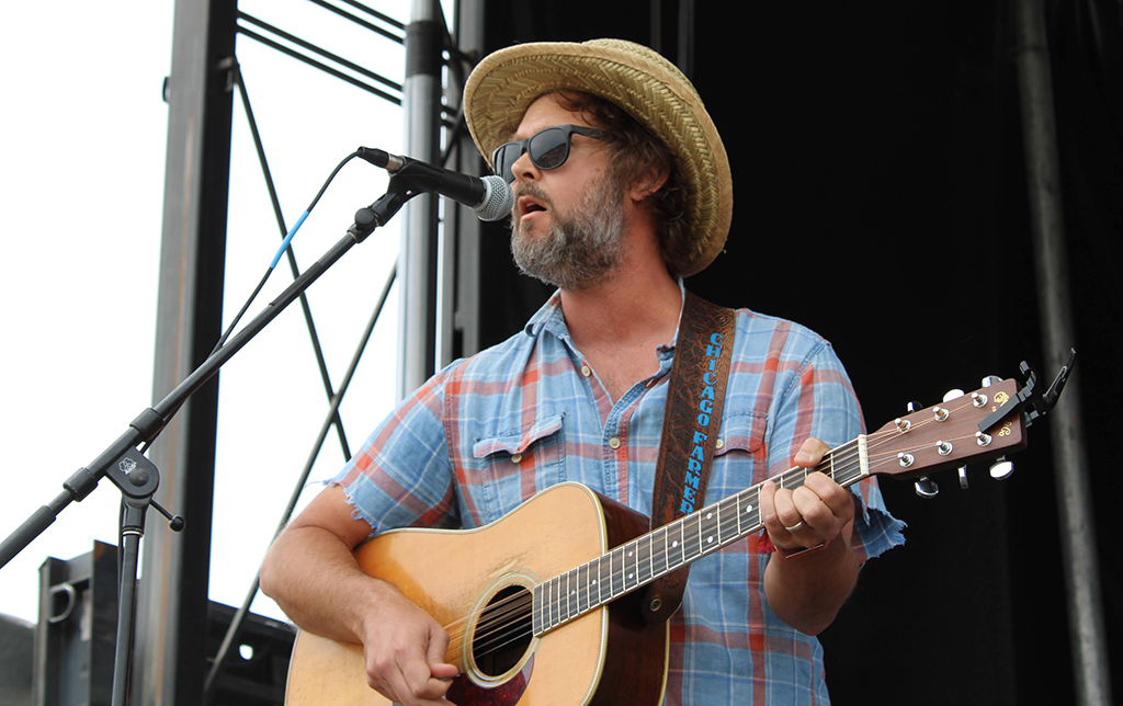 A man playing an acoustic guitar on a stage. He is wearing a blue plaid shirt and a straw hat. The guitar is light in color with a dark neck. The man is singing into a microphone on a stand. The background is a black stage with metal scaffolding.