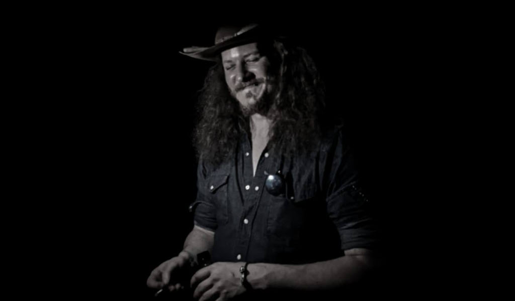 The image is a black and white photo of a person with long curly hair wearing a cowboy hat and a button-down shirt. They are standing against a black background. The person is wearing a black cowboy hat with a silver band and a black button-down shirt with silver buttons. Their hair is long and curly, falling over their shoulders.