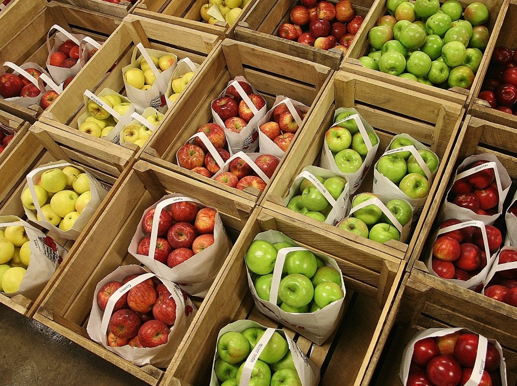 Crates of white bags full of apples are sorted into rows by color. They are in vertical lines starting with red apples, yellow, red, green, and red apples.