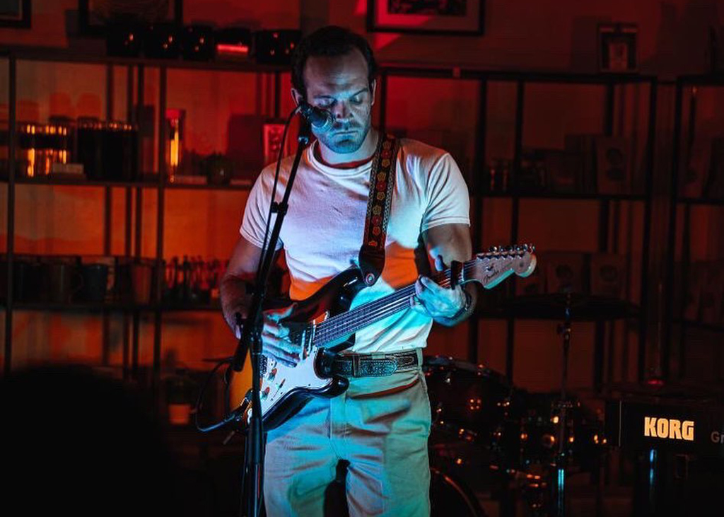 The image shows a musician playing a guitar on a stage. The musician is wearing a white t-shirt and blue jeans, and the guitar is black and white with a strap. In the background, there is a drum set and a keyboard with the brand “KORG” visible. There are also shelves with bottles and glasses. The lighting is dim with a red hue.