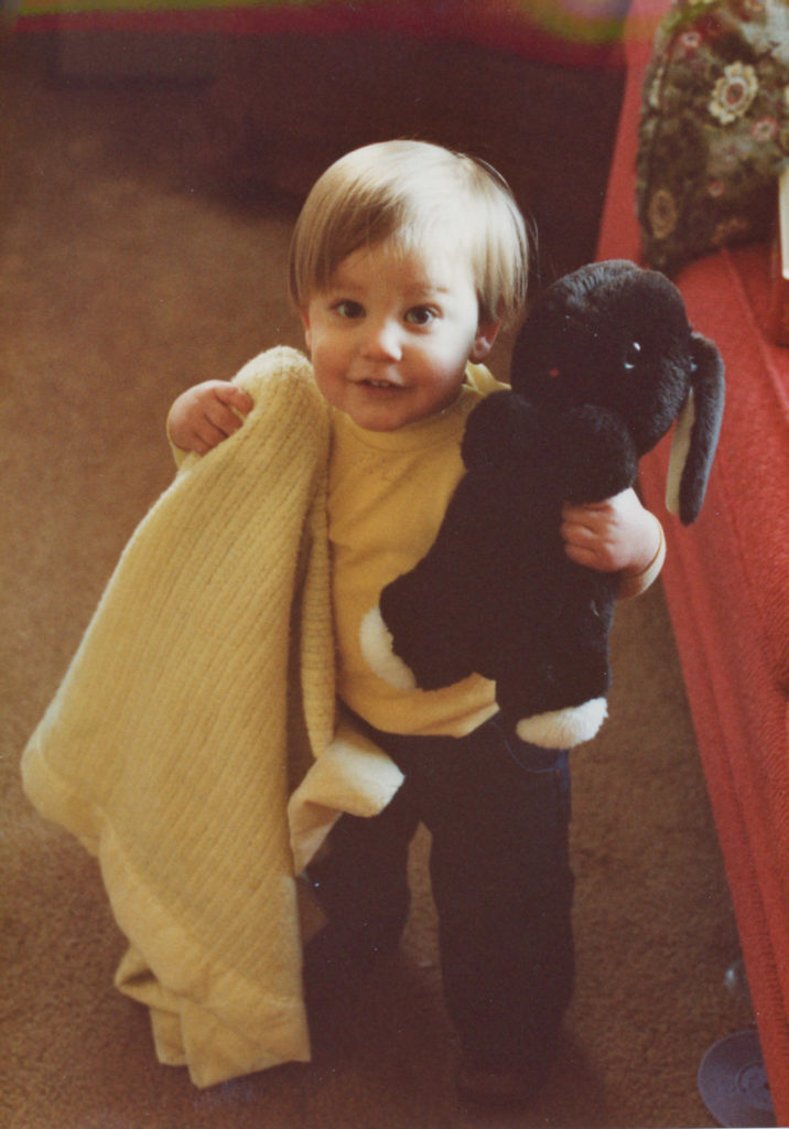Grant Thomas as a toddler. He has blonde hair, is carrying a blanket and a large black stuffed bunny. 