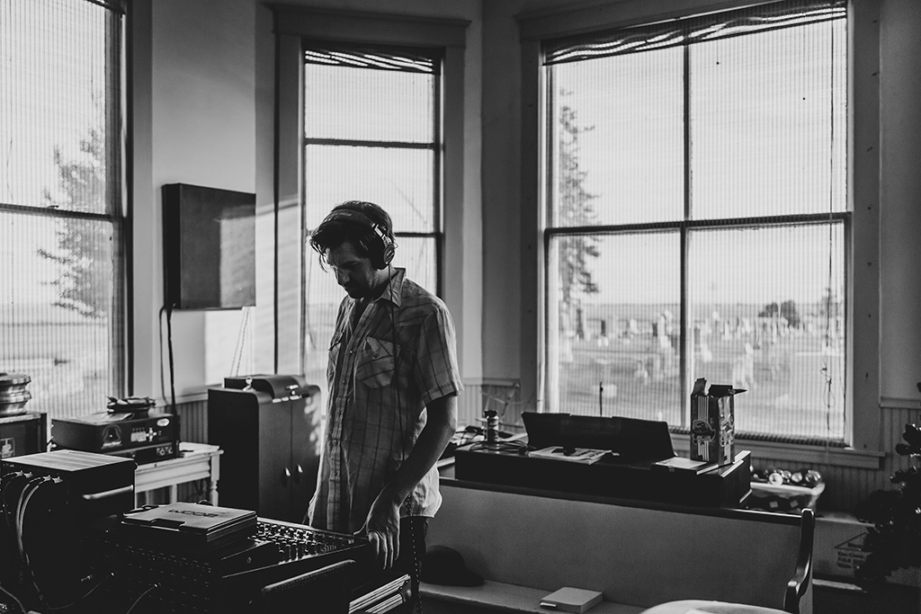This is a black and white photo of a person in a room with a window. The person is wearing a plaid shirt and headphones. The person’s face is blurred out with a white rectangle. The room has a desk with a computer and other equipment on it. The window has blinds and you can see trees outside. The photo has a moody and contemplative feel to it.