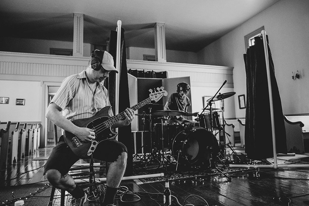 This is a black and white photo of two musicians performing in a room with wooden floors and white walls. The musician on the left is playing a bass guitar and is wearing a baseball cap and a striped shirt. The musician on the right is playing the drums and is wearing a beanie and a tank top. There are microphone stands and cables scattered around the floor. The room has a window with curtains on the right side and a door on the left side. The photo has a grainy texture, giving it a vintage feel.