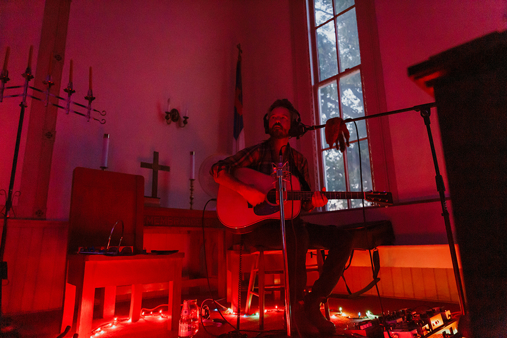 This is a photo of a musician playing guitar in a dimly lit church. The musician is sitting on a stool and is wearing a hat. The musician is playing an acoustic guitar and is singing into a microphone. The musician is surrounded by red string lights and music equipment. The background consists of a stained glass window and a cross on the wall. The mood of the photo is intimate and cozy.