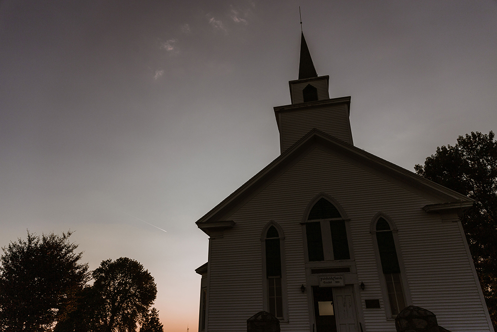 This is a photo realistic image of a white church with a steeple at dusk. The church is a traditional New England style with a pointed steeple and a cross on top. The sky is a dark orange color with a few clouds and a jet trail. The church is surrounded by trees and there is a street lamp in the foreground. The image is taken from a low angle, looking up at the church.