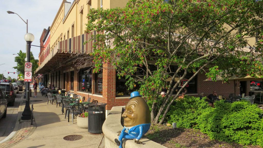 A street corner with a brick building with striped awnings and plastic tables and chairs along the sidewalk. There is a landscaped area with bushes and a small tree, and a statue of an egg dressed in a blue suit.