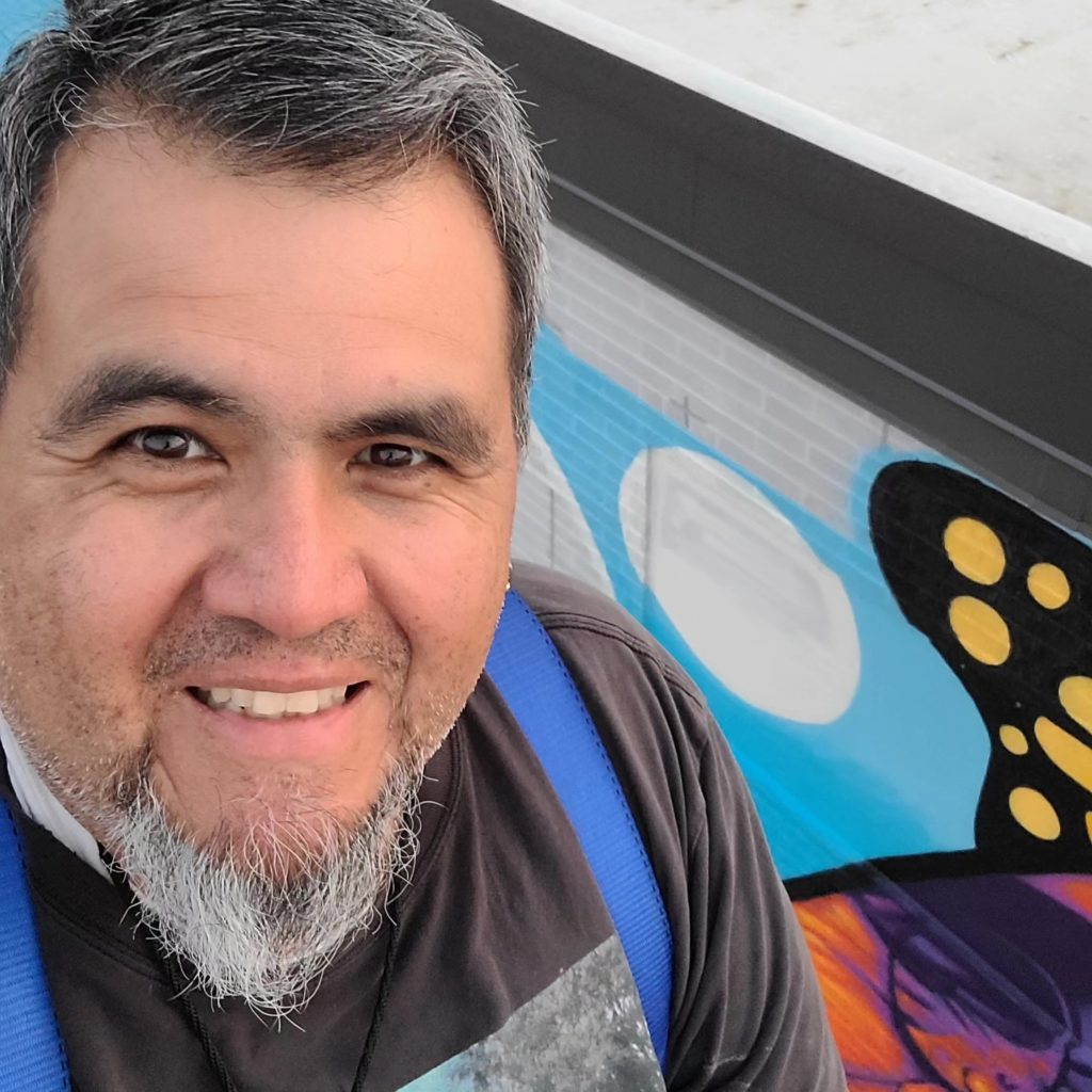 a close up shot of Jose Vazquez standing in front of the CUPHD mural. He is smiling at the camera; he has black and grey hair and a beard. 