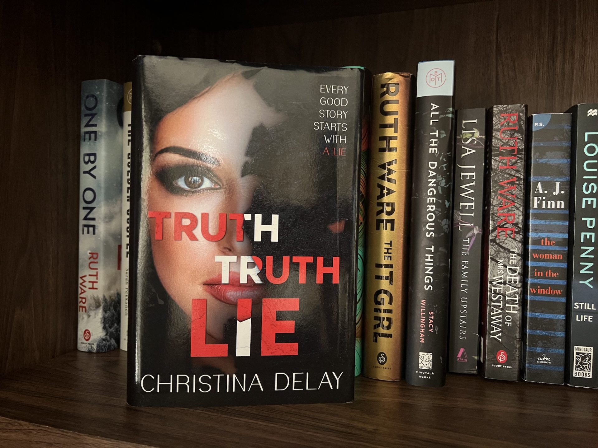 Christina Delay's Truth Truth Lie sits on on a bookshelf with other thrillers behind it. The cover shows a beautiful woman's face half covered with the title large in bold red letters