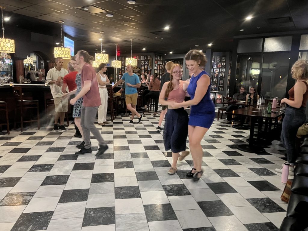 Couples dance lindy hop at Hamilton Walker's. The floor is black and grey black checkered. There are several couples on the floor, mostly dressed casually. The bar is visible in the background.