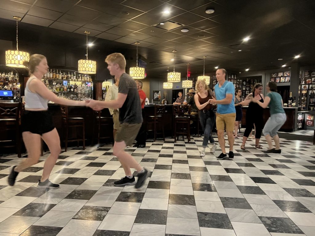 Couples dance lindy hop at Hamilton Walker's. The floor is black and grey black checkered. There are several couples on the floor, mostly dressed casually. The bar is visible in the background.