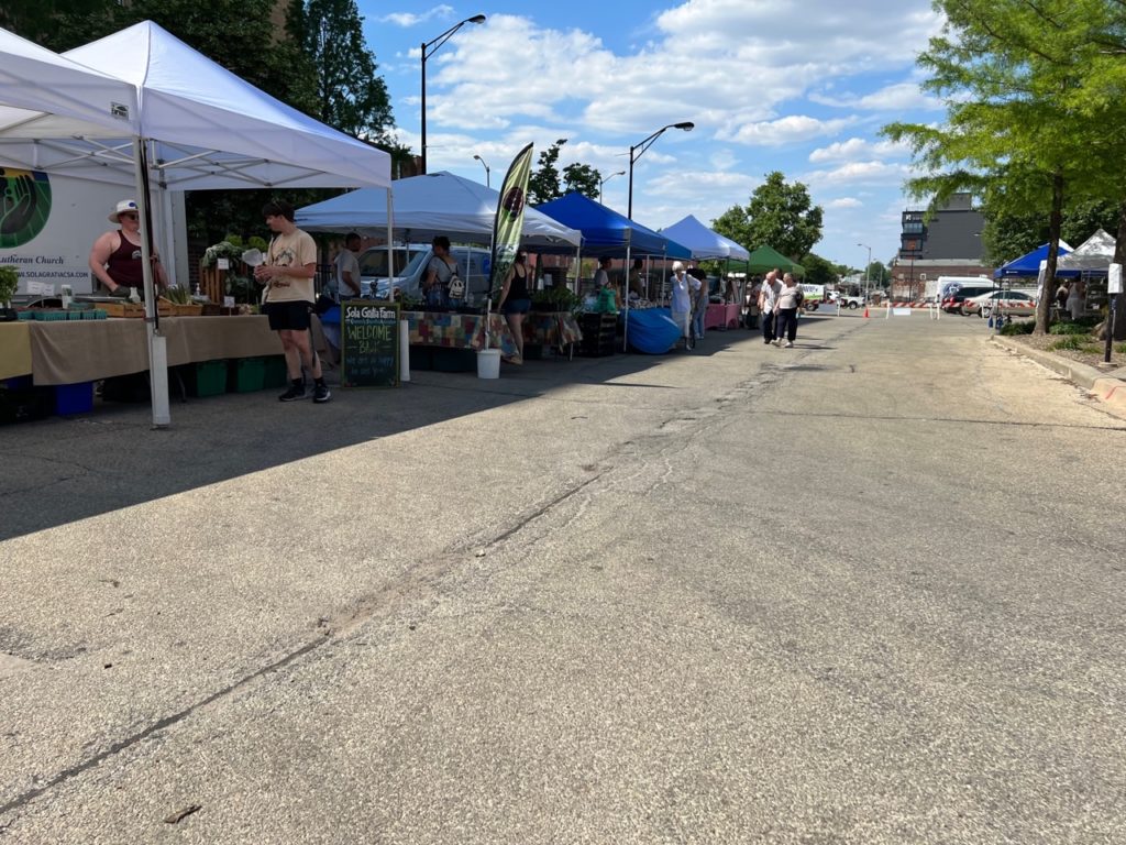 The outdoor Champaign farmers' market has farms with white and blue tents and a few shoppers under a blue sky with fluffy clouds in Downtown Champaign. Photo by Alyssa Buckley.