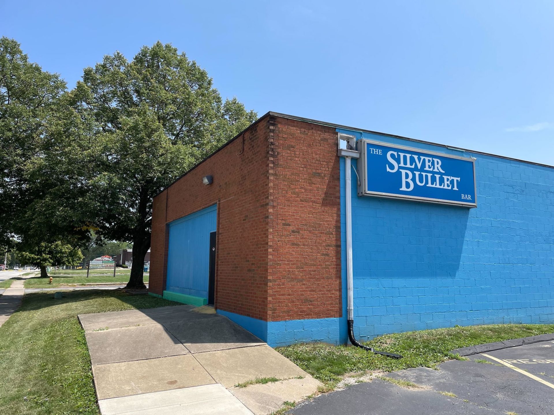 Photo of Silver Bullet in Urbana. Building has a brick front with a bright blue side wall and front panel. There is a blue sky and trees in the background.