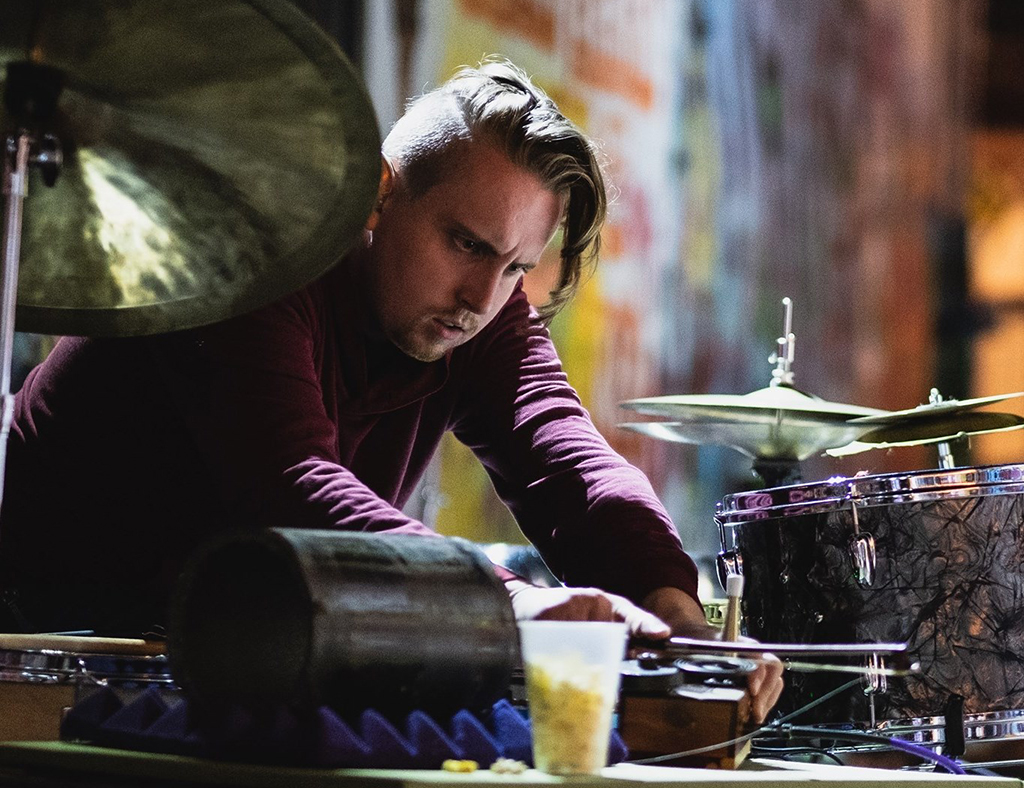 This photo is a photo-realistic image of a drummer playing a drum set in a dimly lit bar. The drummer is wearing a maroon shirt and is playing a drum set with a snare drum, cymbals, and a hi-hat. There is a drink in a plastic cup on the drum set. The background consists of a brick wall with graffiti on it and a cymbal.