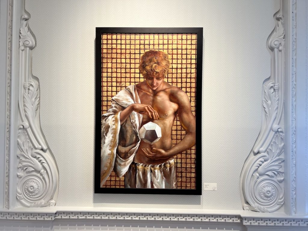 Zeus is pictured shirtless with fabric hanging over one shoulder, He has unruly blonde hair, he is looking down at a silver ball suspended between his hands. The background is made up of tiny gold squares