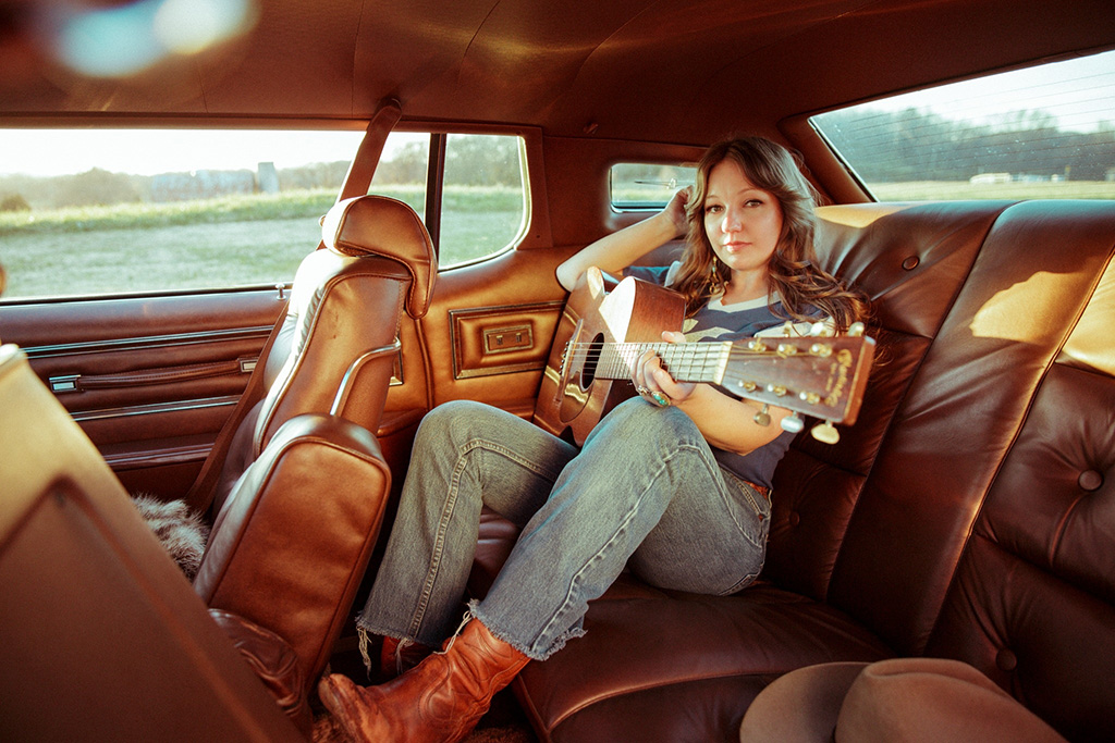 Woman holding a guitar in the back of a car completely upholstered in leather.
