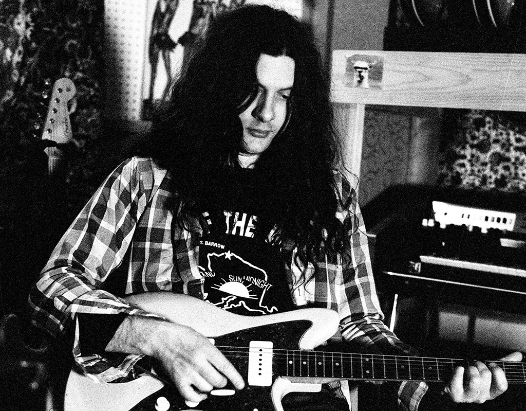 A black and white photo of a man with long hair, sitting down and playing guitar while looking down at the ground.