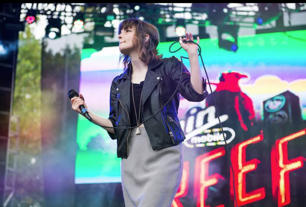A white girl with shoulder length light brown hair holds a microphone and a mic cord onstage while looking up into the crowd. There is a large neon advertising sign on the stage behind her.