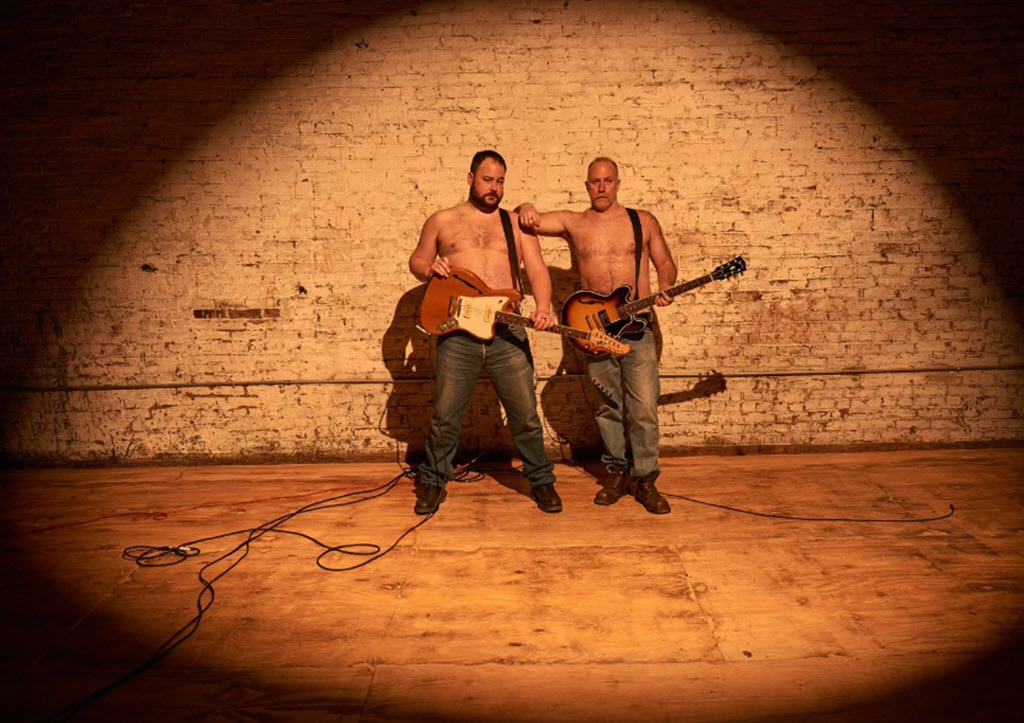 Two white men shirtless with blue jeans and wearing guitars stand closely to one another on a stage with a brick backdrop.