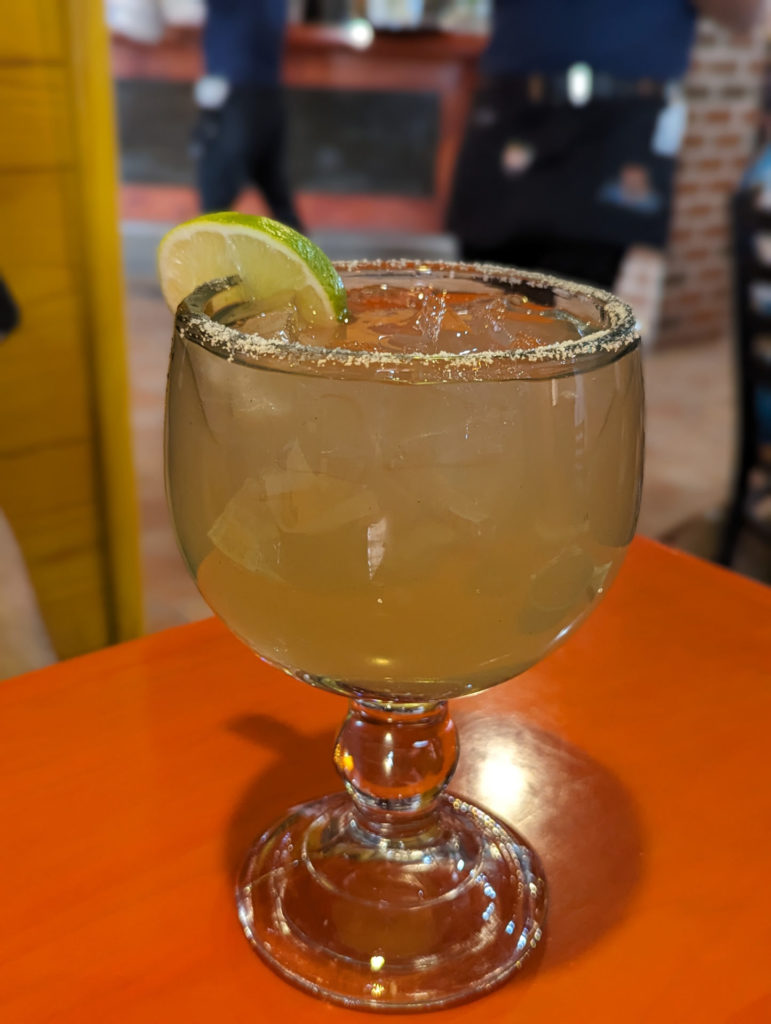 A large margarita glass filled with an iced lime margarita with a salted rim and a lime wedge on top of an orange table. Photo by Tayler Neumann.
