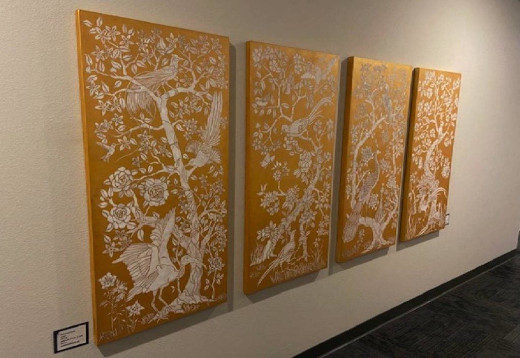 A group of four vertical rectangular paintings hang on a wall. They all have a gold background white white trees and birds taking up space across the canvases. 