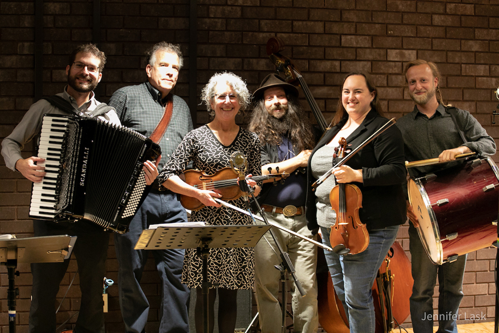 This photo shows a group of musicians playing various instruments in front of a brick wall. The instruments being played include an accordion, a violin, a guitar, and a drum. There is a music stand in front of the violinist.