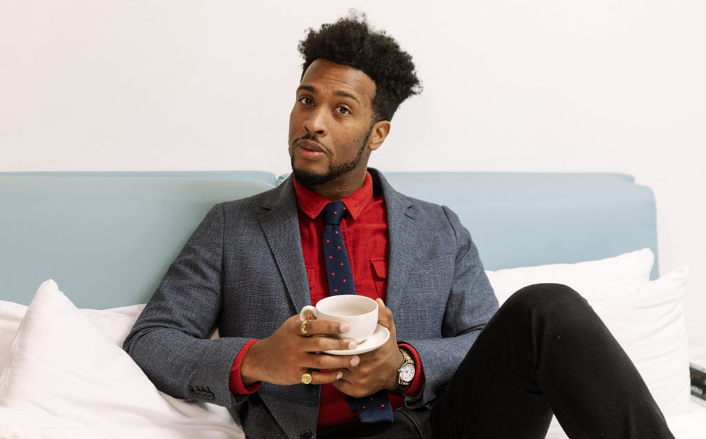 This photo is a photo-realistic image of a person sitting on a blue couch with a white pillow. The person is holding a white coffee cup and saucer in their hands. They are wearing a gray blazer, red shirt, and black pants. The background is a white wall.