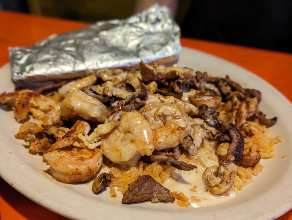 Big sauteed shrimp, steak and chicken pieces on top of rice with foil-wrapped tortilla in the back ground on top of a cream colored plate. Photo by Tayler Neumann.