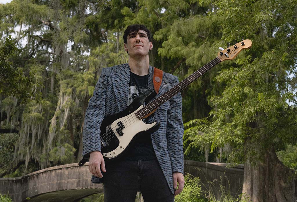 This is a photo realistic image of a person holding a white electric guitar in a park. The person is wearing a blue and white checkered blazer and a black t-shirt. The guitar is white with a black pickguard and a maple neck. The background consists of a park with trees and a stone bridge.