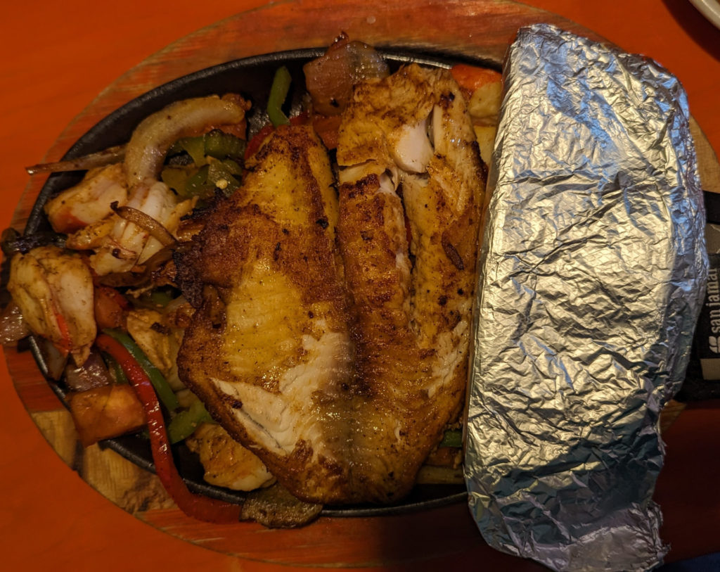 A top view of a cast iron plate with a piece of grilled fish on top of shrimp, onions, and peppers with foil-wrapped tortillas on the left side. Photo by Tayler Neumann.