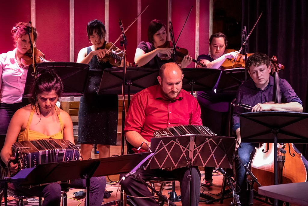 This is a photo realistic image of a group of musicians playing on a stage. The musicians are playing various instruments such as violins, cellos, and accordions. The musicians are seated on chairs and are reading music from music stands. The background consists of a red curtain and stage lighting