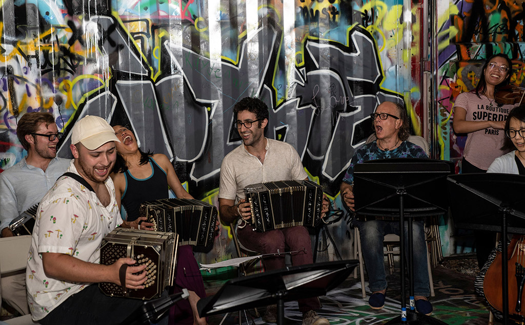 This is a photo of a group of musicians playing in front of a colorful graffiti wall. The musicians are playing various instruments, including accordions, a guitar, and a drum set. The musicians are sitting on a green carpeted floor. The graffiti wall is covered in various colors and designs, including letters and abstract shapes.