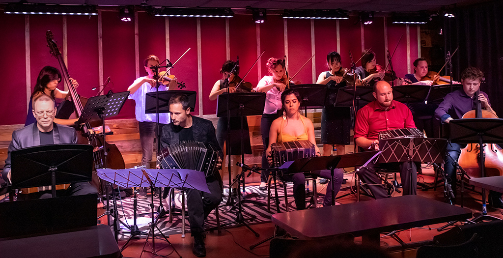 This is a photo realistic image of a group of musicians playing string instruments on a stage. The stage has a red curtain in the background and is lit by stage lights. The musicians are seated on black chairs and are playing violins, violas, cellos, and double basses. The musicians are using music stands to hold their sheet music. The foreground of the image shows empty chairs and tables, suggesting that this is a performance venue.